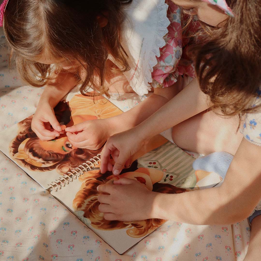 Twee kinderen die stickers plakken op de ogen van de geïllustreerde meisjes in het open stickerboek.