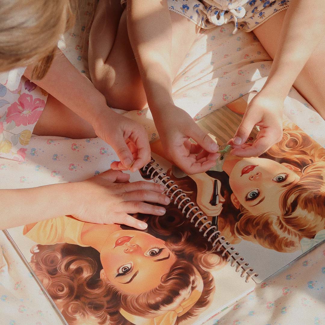 Twee kinderhanden die stickers plakken op de geïllustreerde gezichten in het open Glam Me Up stickerboek.