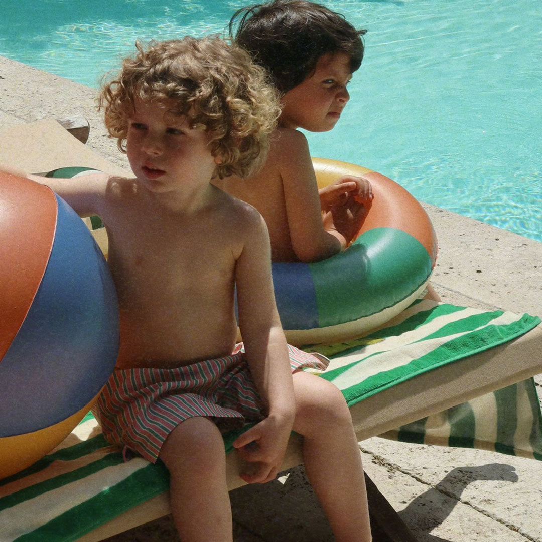 Twee kinderen zitten op een ligstoel met het Konges Sløjd gestreepte strandlaken bij een zwembad.