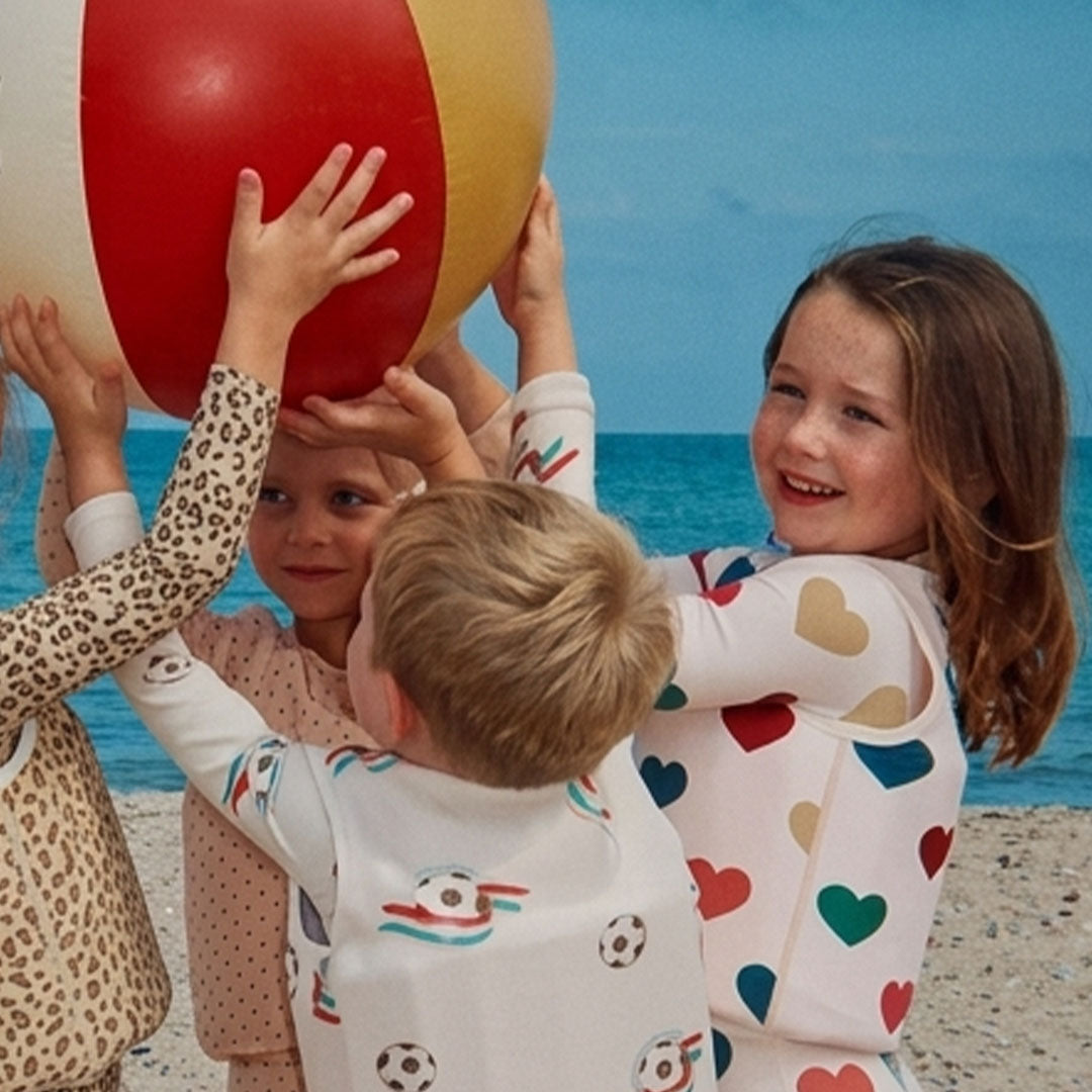 Meerdere kinderen spelen met een strandbal op het strand, één kind draagt het Konges Sløjd zwemvest met hartenpatroon.