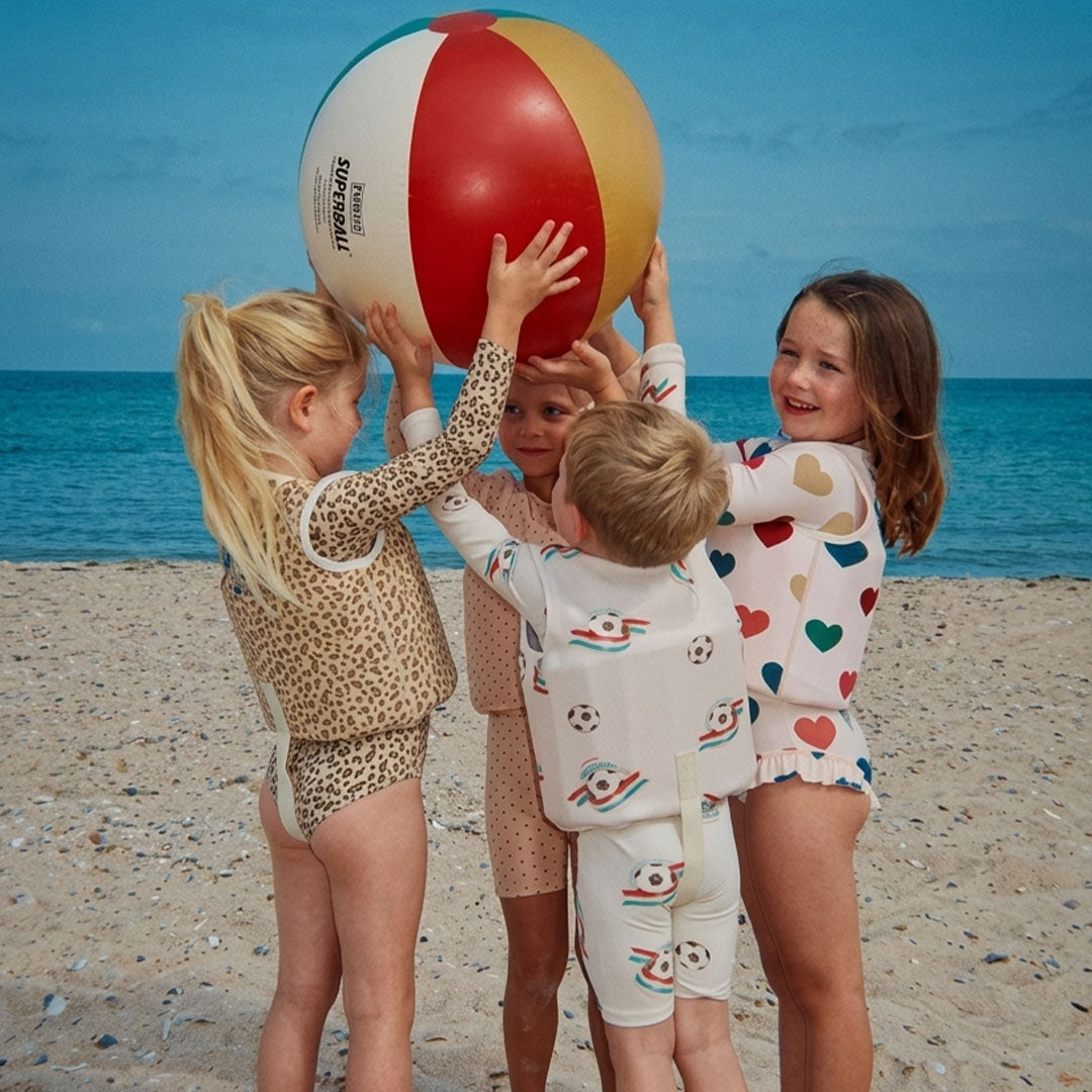Vier kinderen op het strand, waarvan één het Konges Sløjd zwemvest met voetbalthema draagt, terwijl ze een grote strandbal vasthouden.