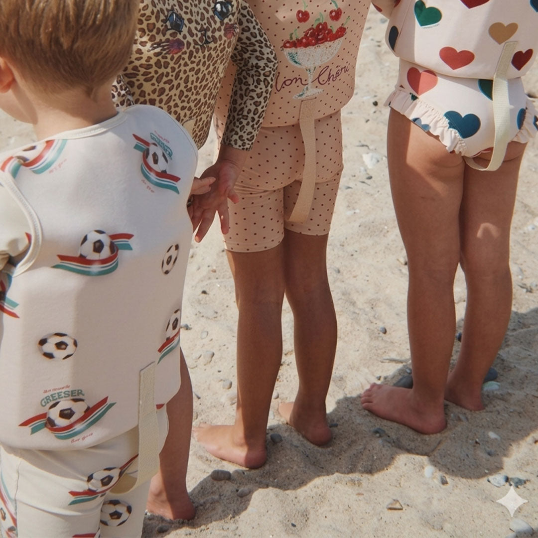 Achterkant van een kind dat het Konges Sløjd zwemvest met voetbalthema draagt, staand op het strand.