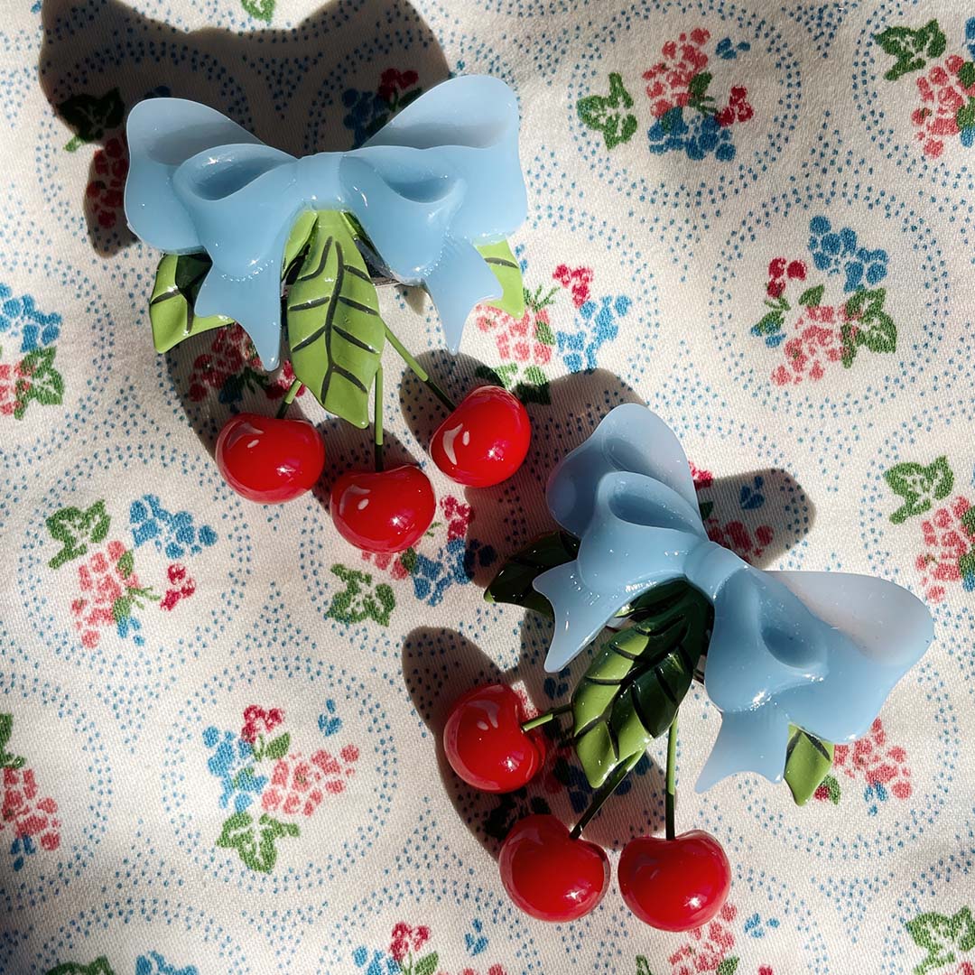 Twee Konges Sløjd kinder haarclips met blauwe strikken en rode kersen liggen op een wit doek met bloemenpatroon.