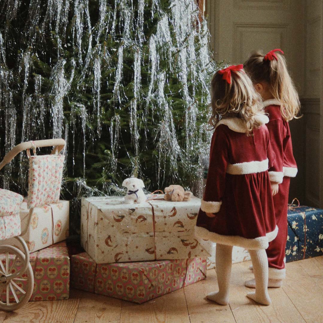 Twee kinderen met rode konges sløjd fluwelen haarclips in het haar, kijkend naar een kerstboom met cadeaus.