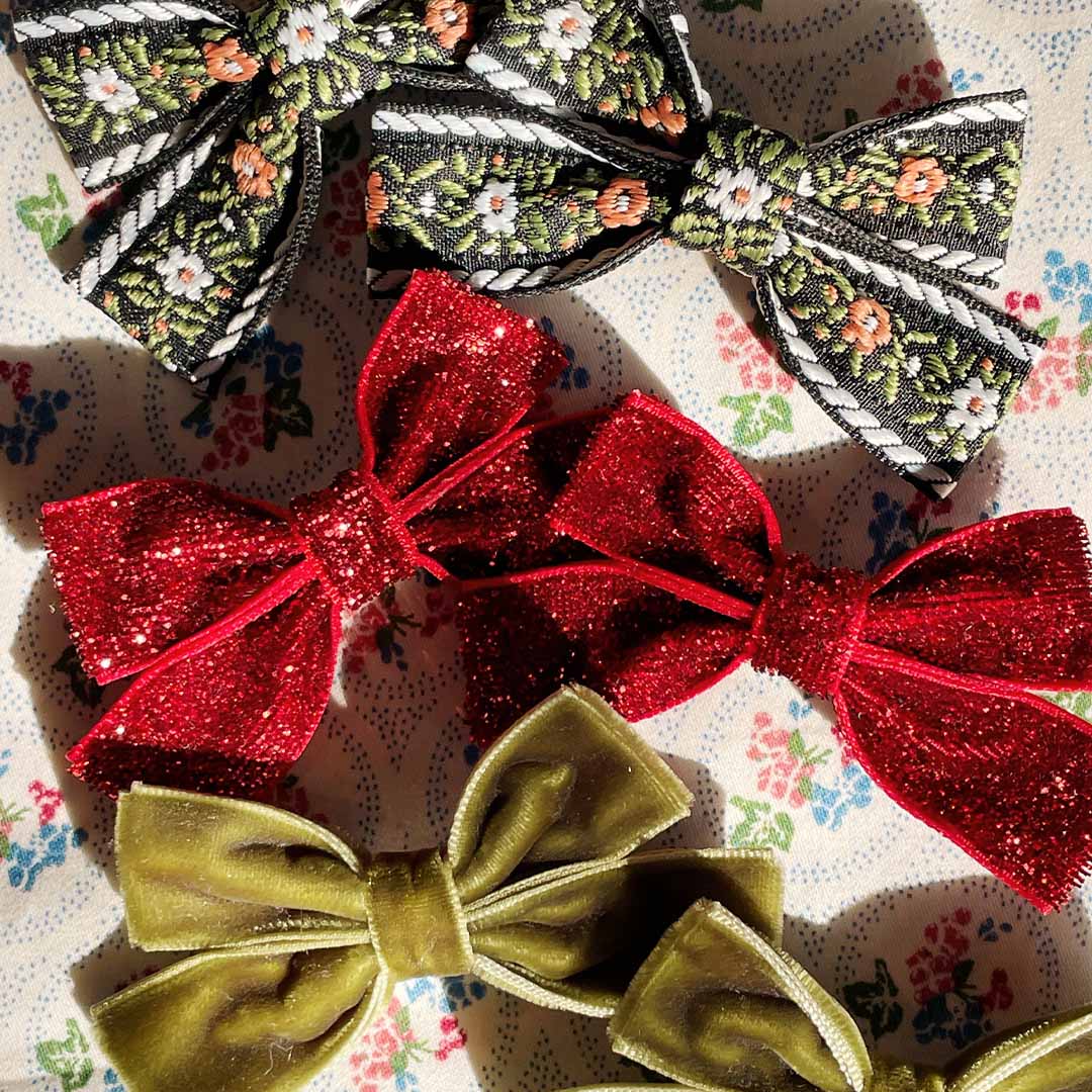 Zes Konges Sløjd haarclips voor kinderen, twee rode glitter, twee zwart met bloemenpatroon en twee olijfgroen fluweel, liggend op een gebloemde stof.