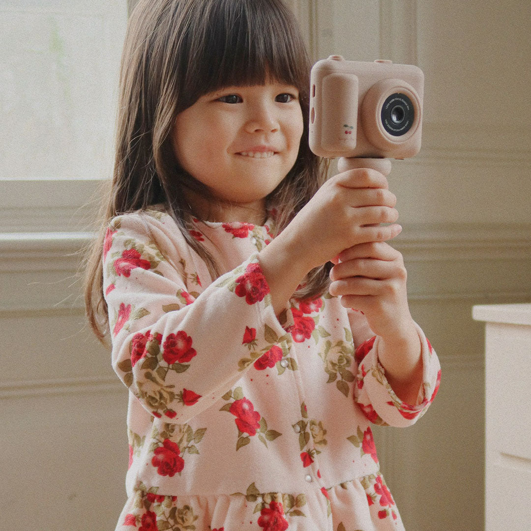 Een lachend meisje in een roze bloemenoutfit houdt de kindercamera vast met het statief als handvat.