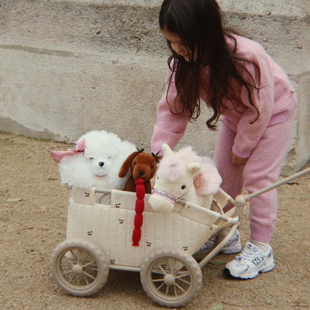 Een kind duwt een beige poppentrekwagen met kersenpatroon waarin drie knuffelhonden zitten.