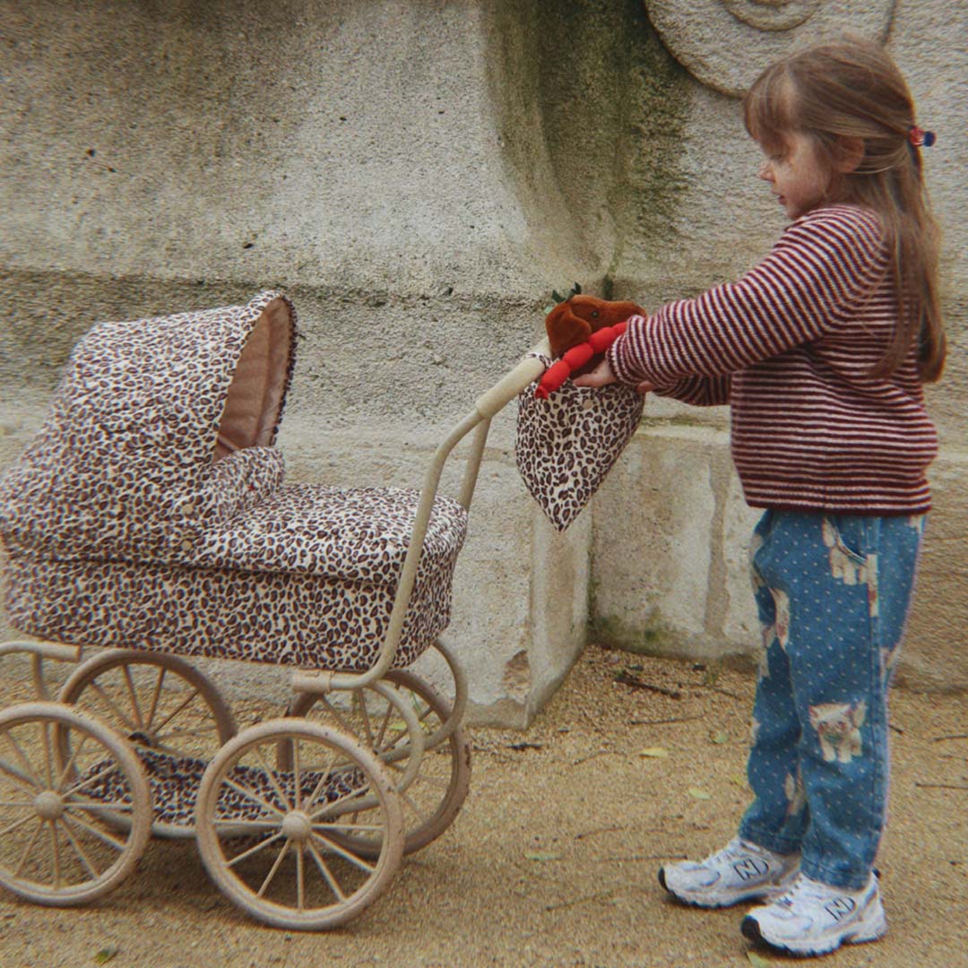 Een kind duwt een Konges Sløjd poppenwagen met leopard print over een pad.