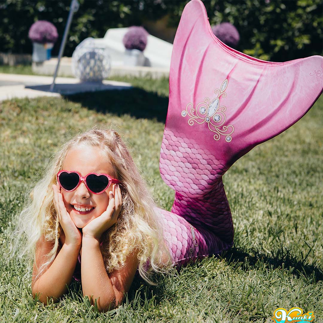 Een meisje met een kuaki mermaids roze zeemeerminstaart en hartjeszonnebril ligt lachend in het gras.