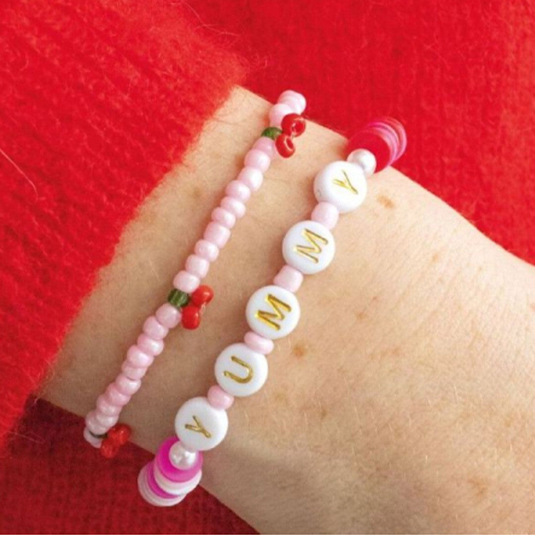 Een arm draagt twee roze DIY armbanden, één met de tekst 'YUMMY' en één met kleine kersjes.