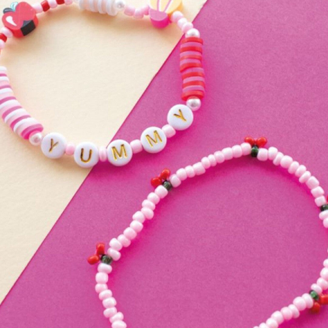 Twee roze DIY armbanden liggen op een roze en beige ondergrond, één met de tekst 'YUMMY' en één met kersjes.
