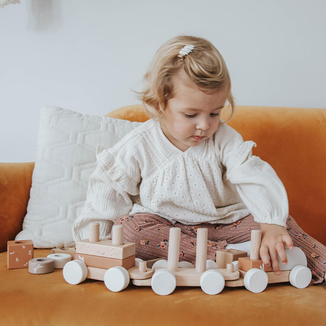Een jong kind zit op een gele bank en speelt met de Label Label houten blokkentrein.