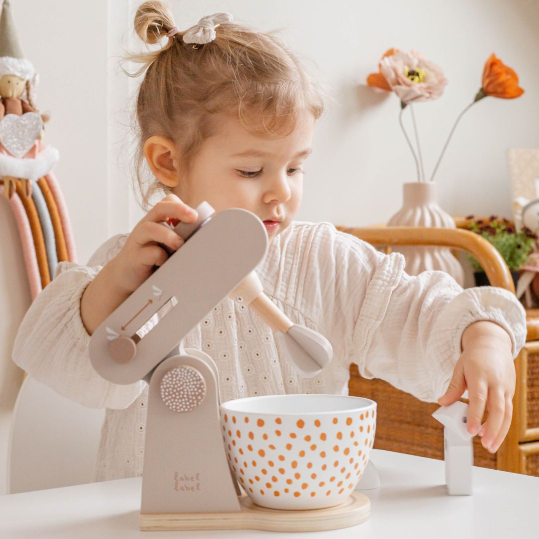 Een kind speelt met de Label Label houten speelgoedkeukenmachine, waarbij de mixerarm omhoog is en ze het melkpak vasthoudt.