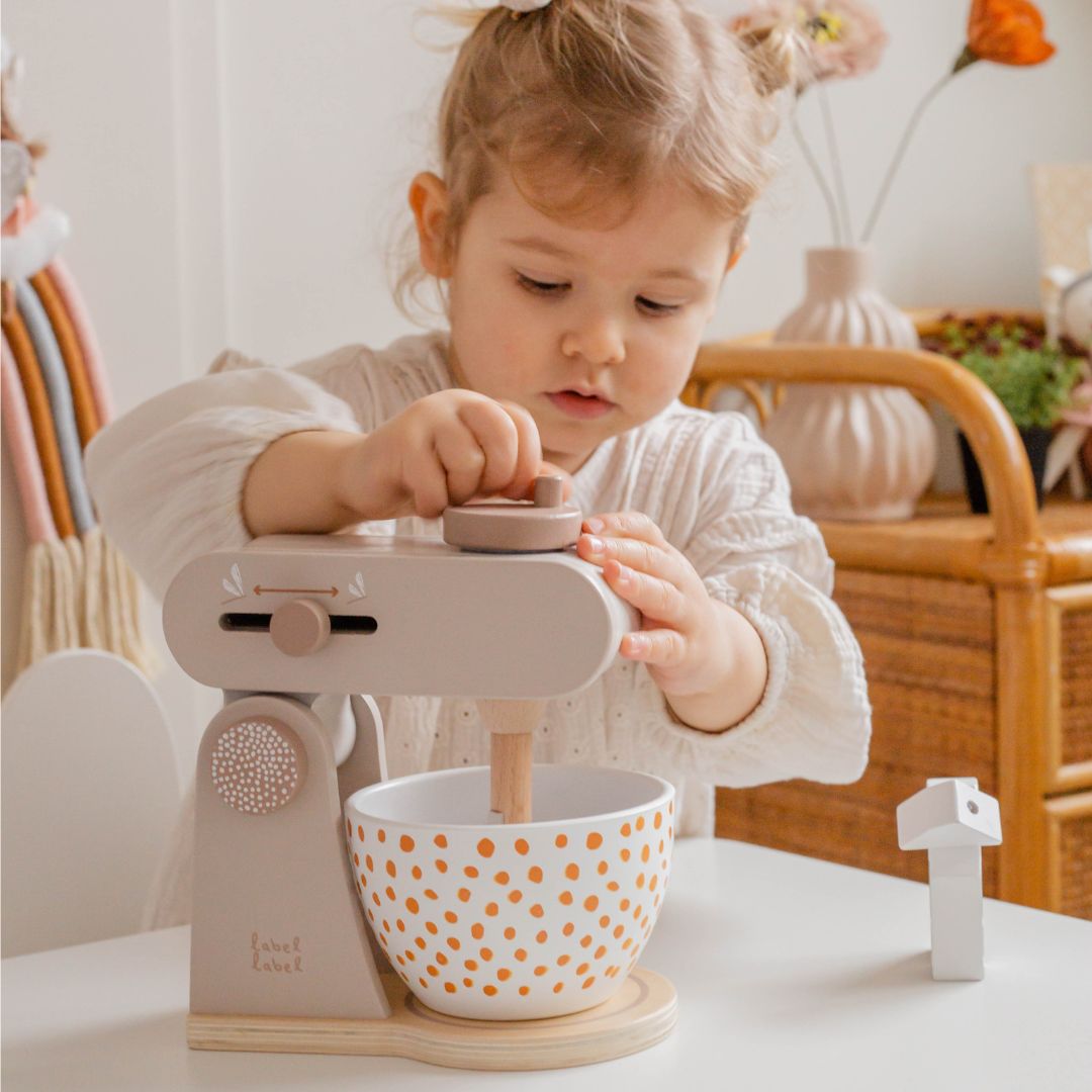 Een kind speelt met de Label Label houten speelgoedkeukenmachine met de mixerarm omlaag en draait aan de knop.