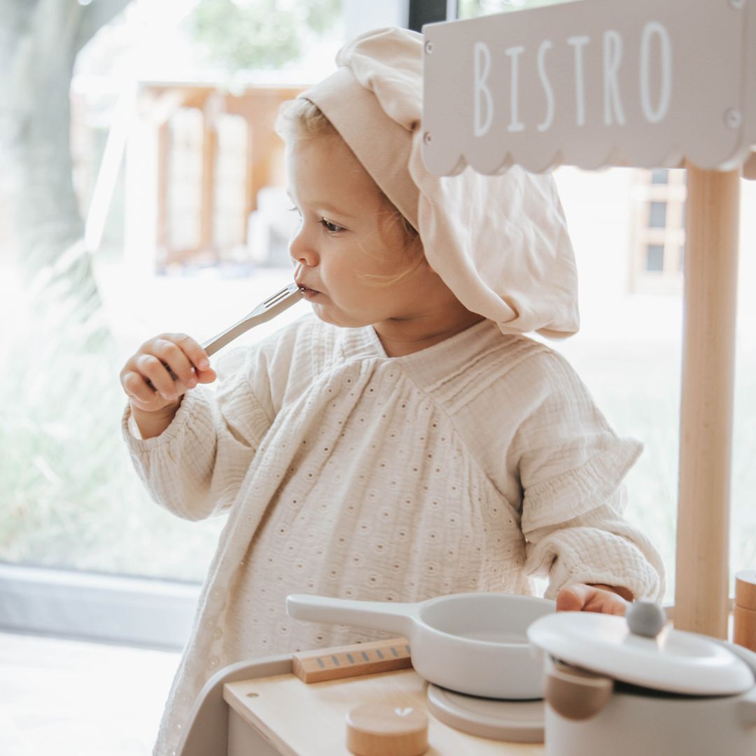 Een kind met koksmuts proeft van een vork bij de Label Label houten kinderkeuken bistro.