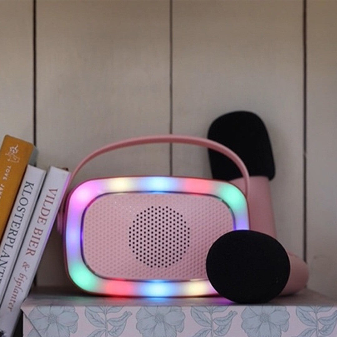 Roze karaokespeaker met regenboogverlichting en een microfoon op een plank naast boeken.