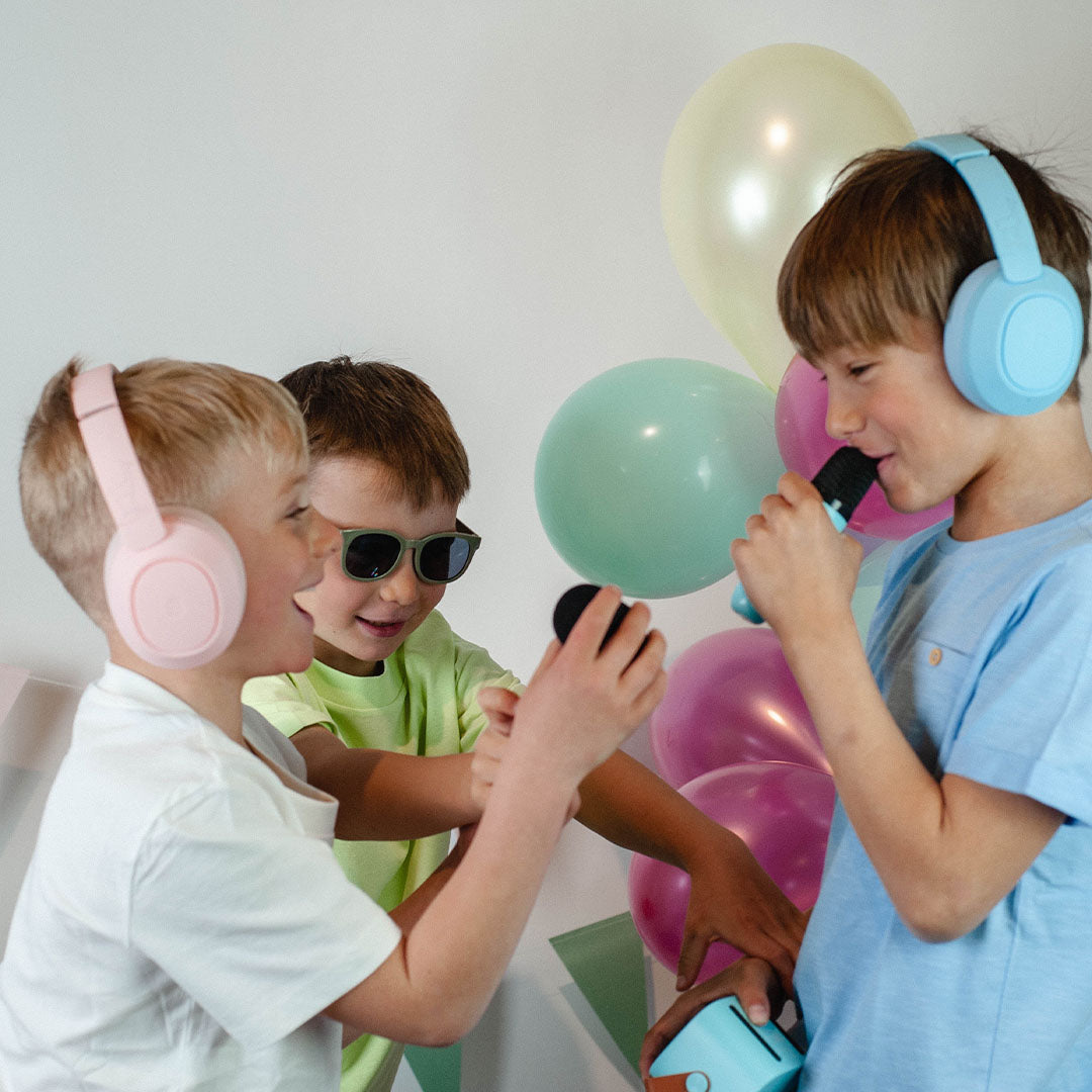 Drie kinderen spelen met microfoons, waarvan één jongen een lichtblauwe lalarma kinderkoptelefoon draagt.