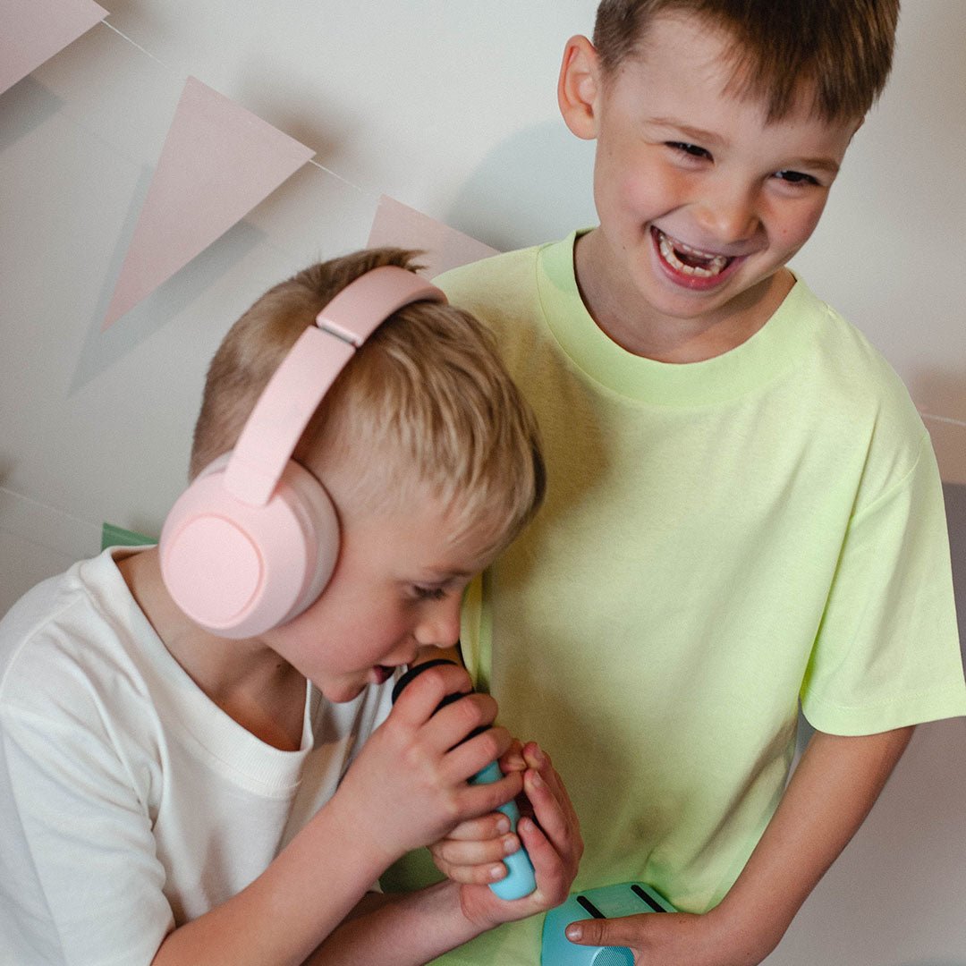 Een jongen met de roze kinderkoptelefoon zingt in een microfoon, terwijl een andere jongen lacht.