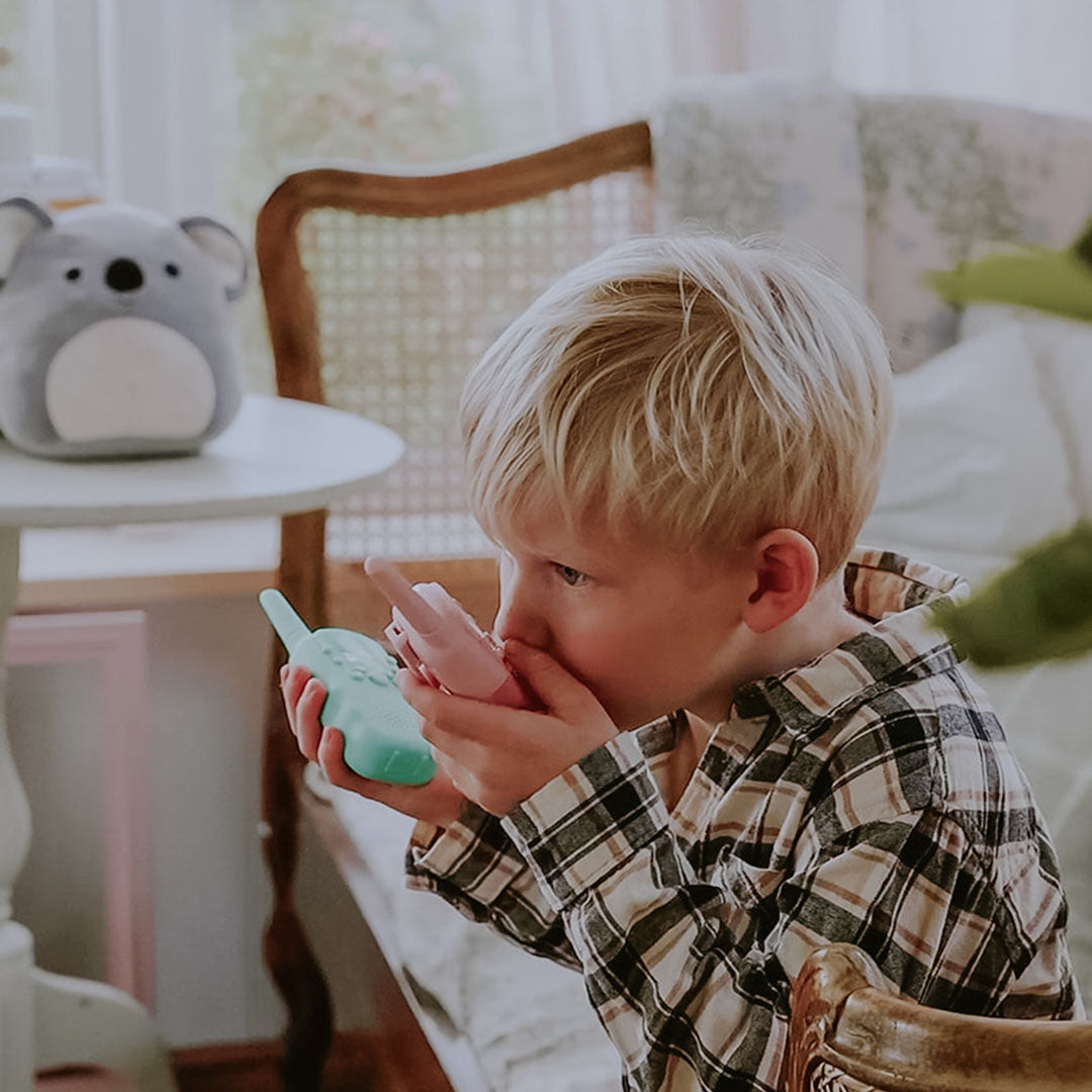 Jonge jongen met blond haar praat in een lalarma copenhagen walkie talkie, met een tweede roze walkie talkie in zijn andere hand.
