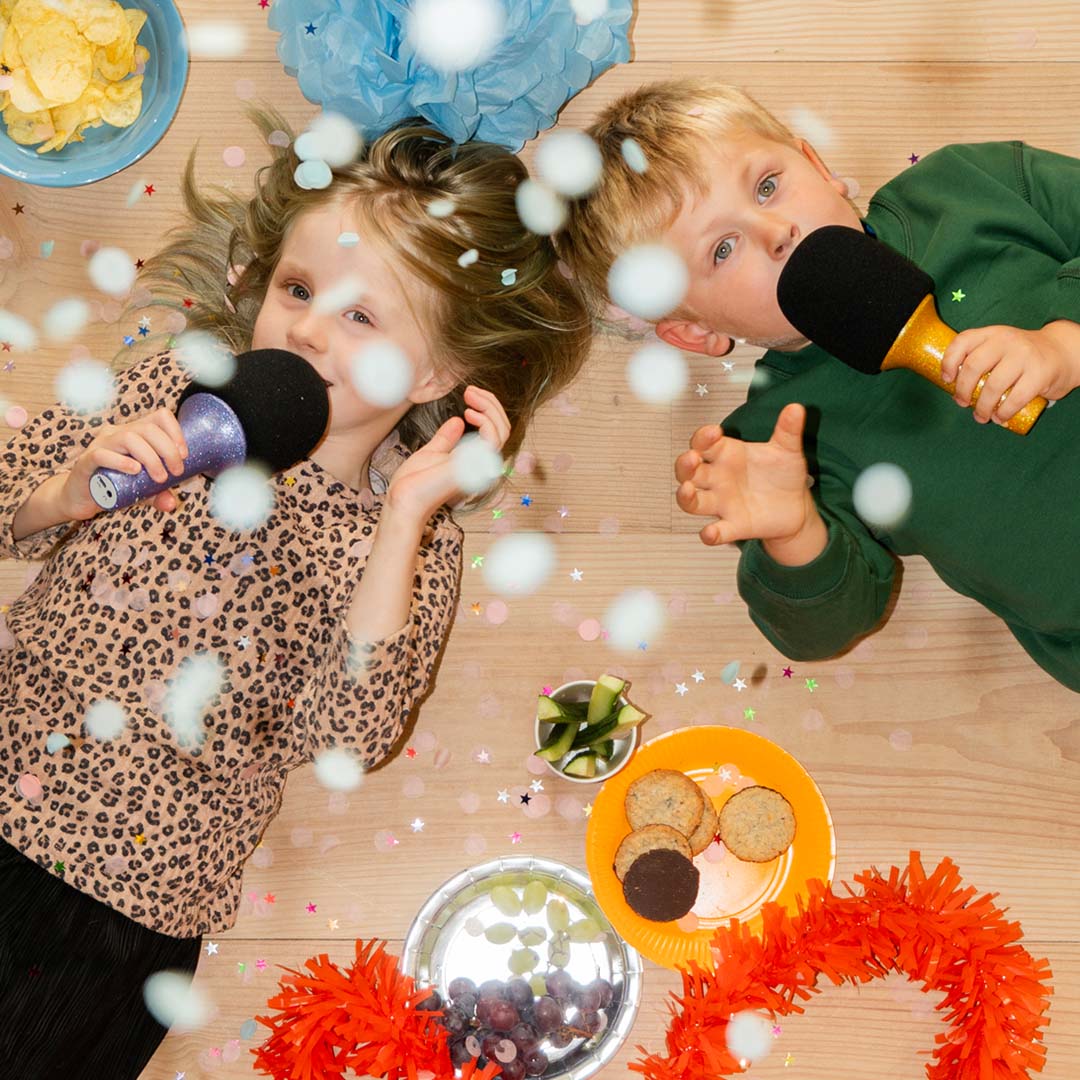 Twee kinderen zingen met glitter karaoke microfoons op een houten vloer vol confetti en snacks.