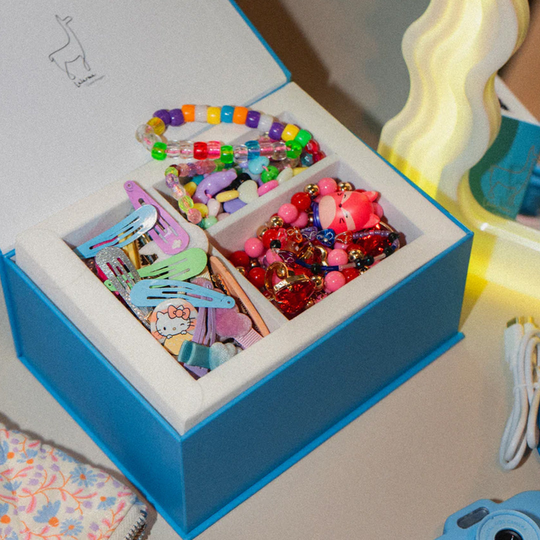 Open blauwe doos met vakken gevuld met kleurrijke kinderaccessoires zoals kralenarmbanden en haarspeldjes.