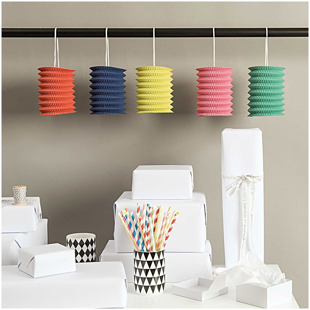 Vijf kleurrijke papieren lampions hangen aan een stang boven een tafel met witte cadeaus en rietjes.