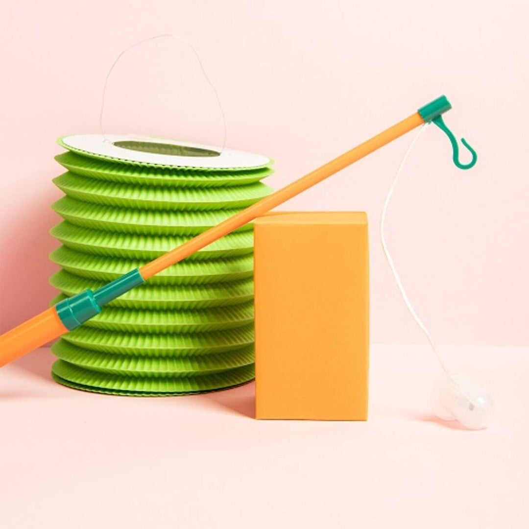 Folat lampionstokje in oranje en groen naast een groene opvouwbare lampion en een oranje blok op een roze achtergrond.