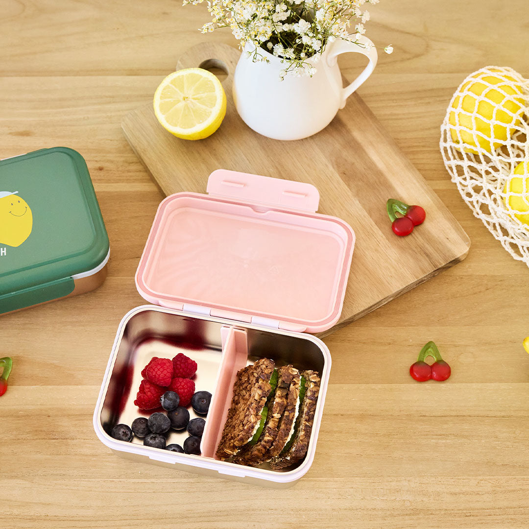 Lässig roze RVS broodtrommel voor kinderen open op een houten tafel, gevuld met fruit en brood.