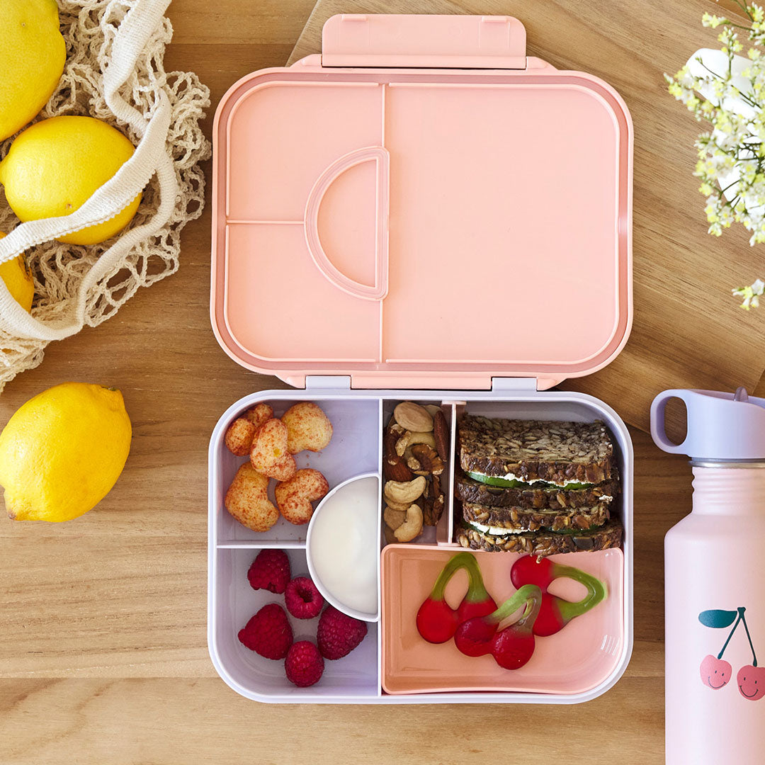 Open roze kinderbroodtrommel met verschillende vakken gevuld met snacks, fruit en een sandwich, naast een roze drinkfles.