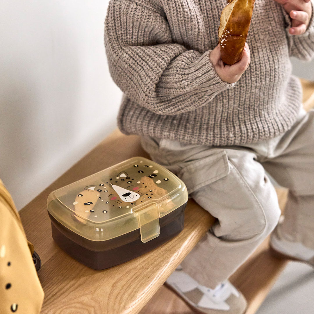 Een kind zit op een houten bankje met de Lässig bento broodtrommel met leopard design naast zich.