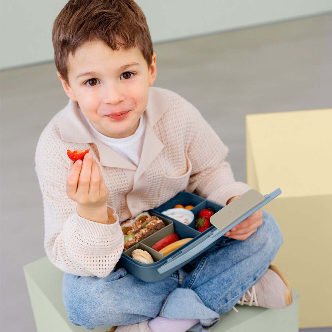 Een lachende jongen eet een aardbei uit de open Lässig kinderbroodtrommel.