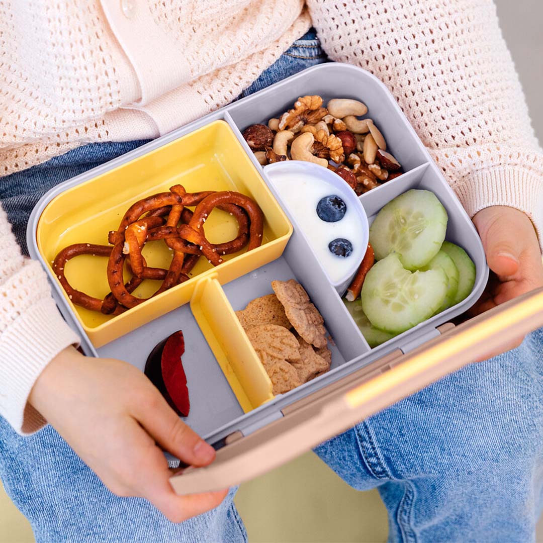 Een open Lässig bento-broodtrommel gevuld met diverse snacks, vastgehouden door kinderhanden.