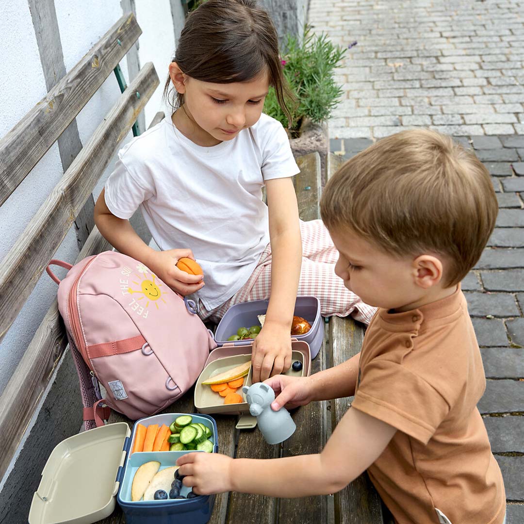 Twee kinderen eten buiten van hun open kinderbroodtrommels met fruit en groenten.