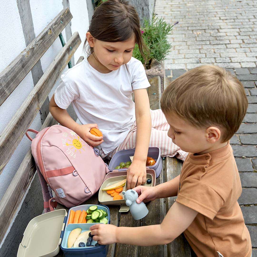 Twee kinderen zitten op een bankje en eten uit twee open Lässig kinderbroodtrommels met verschillende snacks.