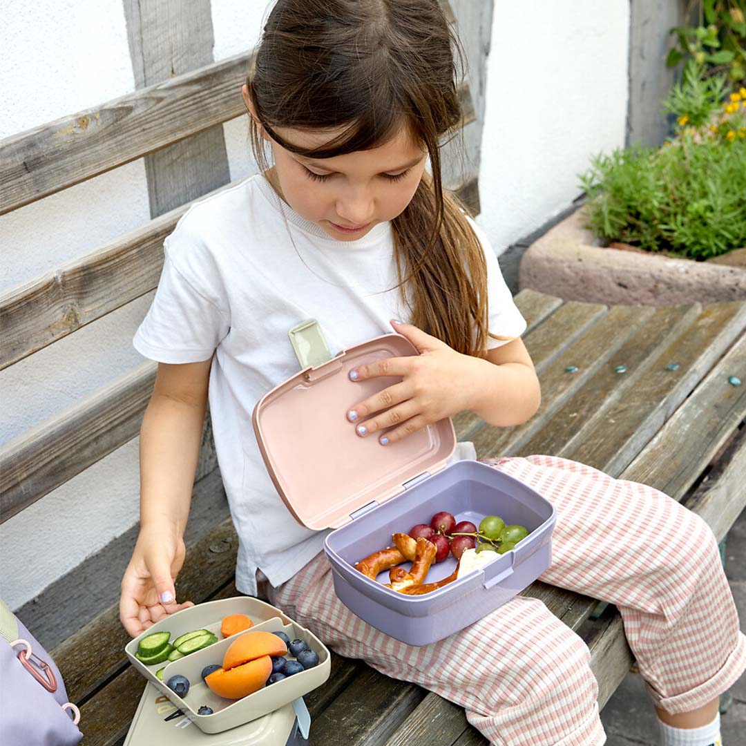 Een kind zit op een bankje en eet uit een open Lässig kinderbroodtrommel gevuld met fruit en snacks.