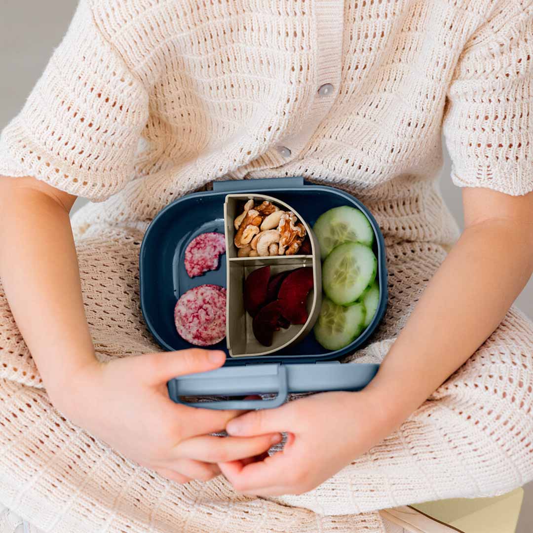 Een kind houdt een open Lässig kinderbroodtrommel vast, gevuld met diverse snacks en met interne verdelers.