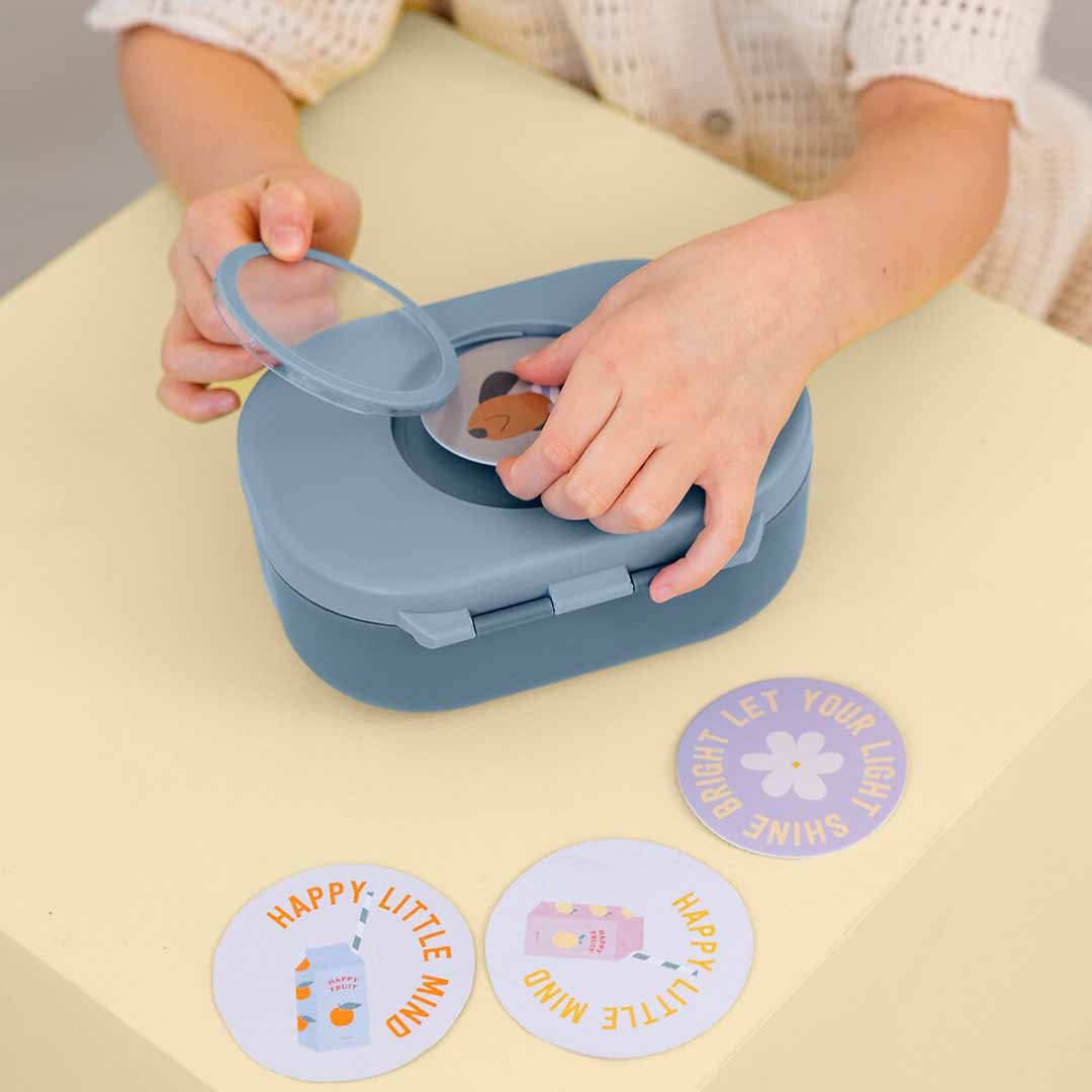 Kinderhanden personaliseren een Lässig kinderbroodtrommel met een ronde sticker van een hondje op de deksel.