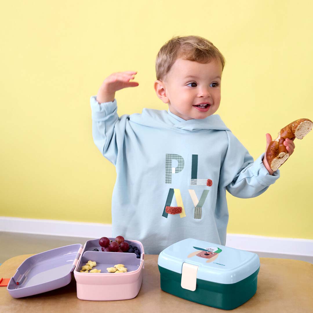 Een peuter met een krakeling bij een open Lässig broodtrommel met snacks en een andere lunchbox op tafel.
