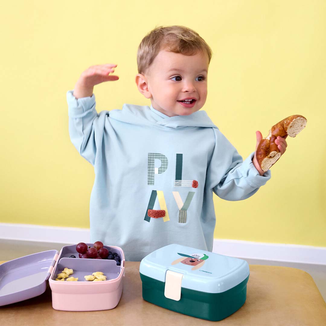 Een kind met een stuk brood naast een open kinderbroodtrommel met hondenthema en een roze lunchbox met fruit.