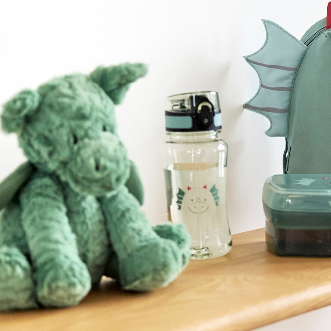 Lässig kinderdrinkfles met draak op een houten plank, naast een groene knuffeldraak en een rugzak met drakenvleugels.
