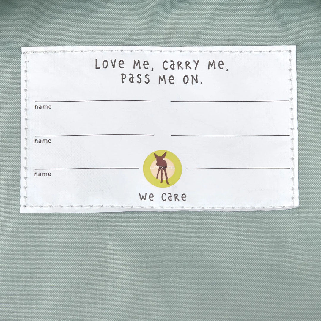 Een wit stoffen label in de rugzak met de tekst 'Love Me, carry Me, pass Me ON' en ruimte voor namen.