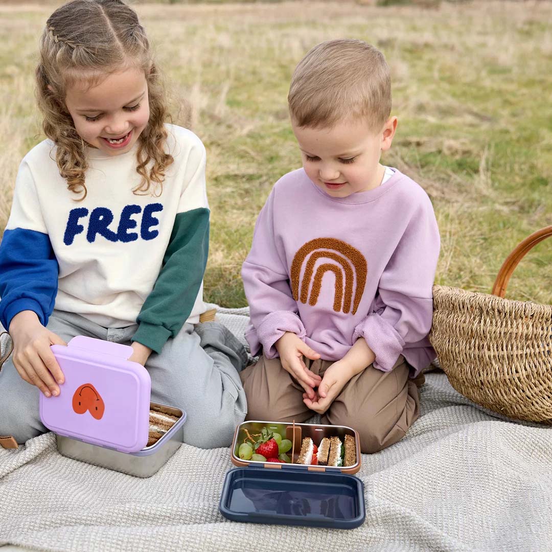Twee kinderen zitten buiten op een deken en openen hun Lässig kinderbroodtrommels tijdens een picknick.