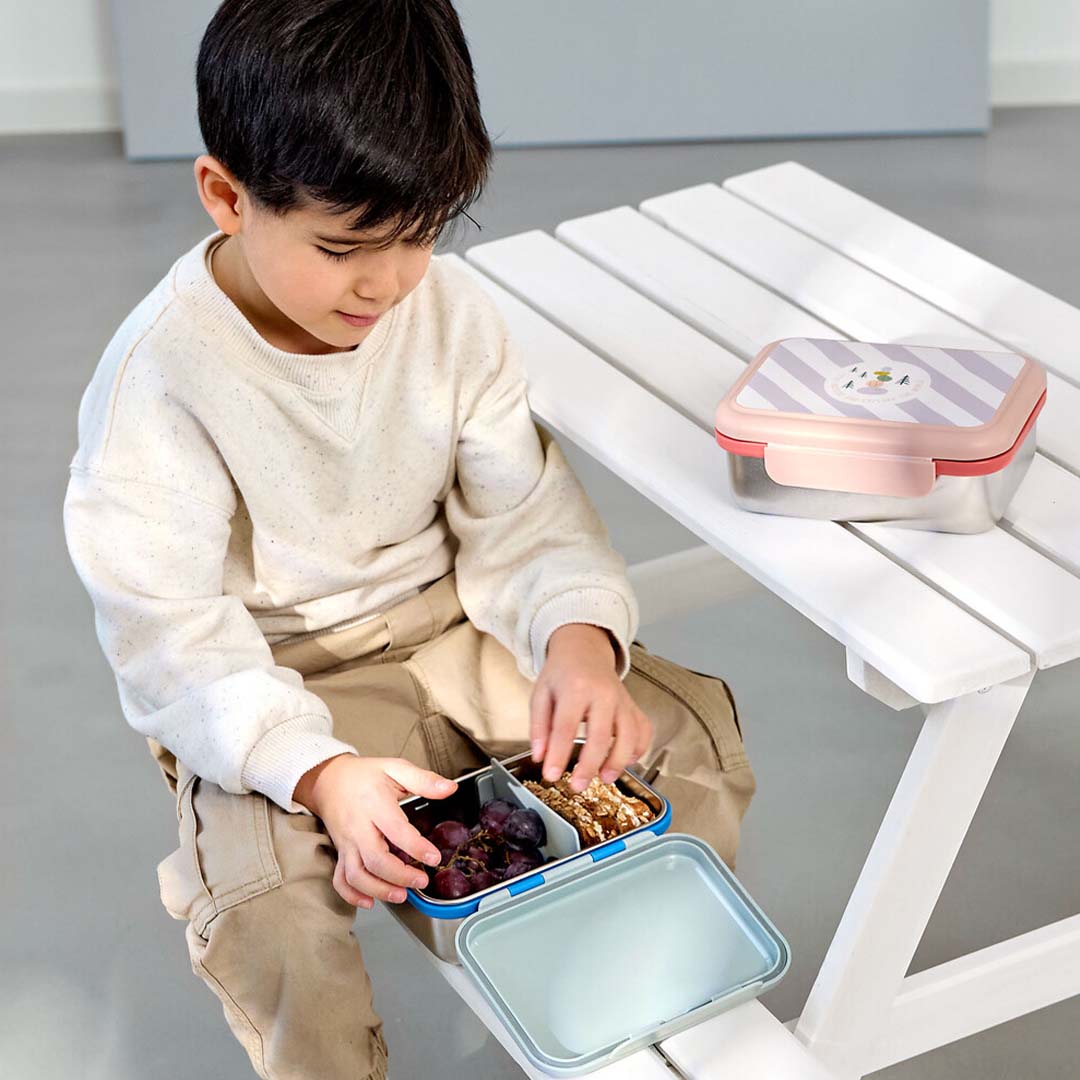 Kind opent een Lässig kinderbroodtrommel met druiven en noten aan een witte picknicktafel.