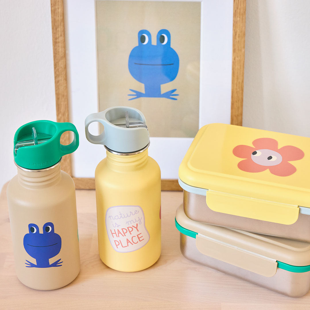 Twee Lässig kinderdrinkflessen en twee lunchboxen op een houten ondergrond, met een kikker en bloem thema.