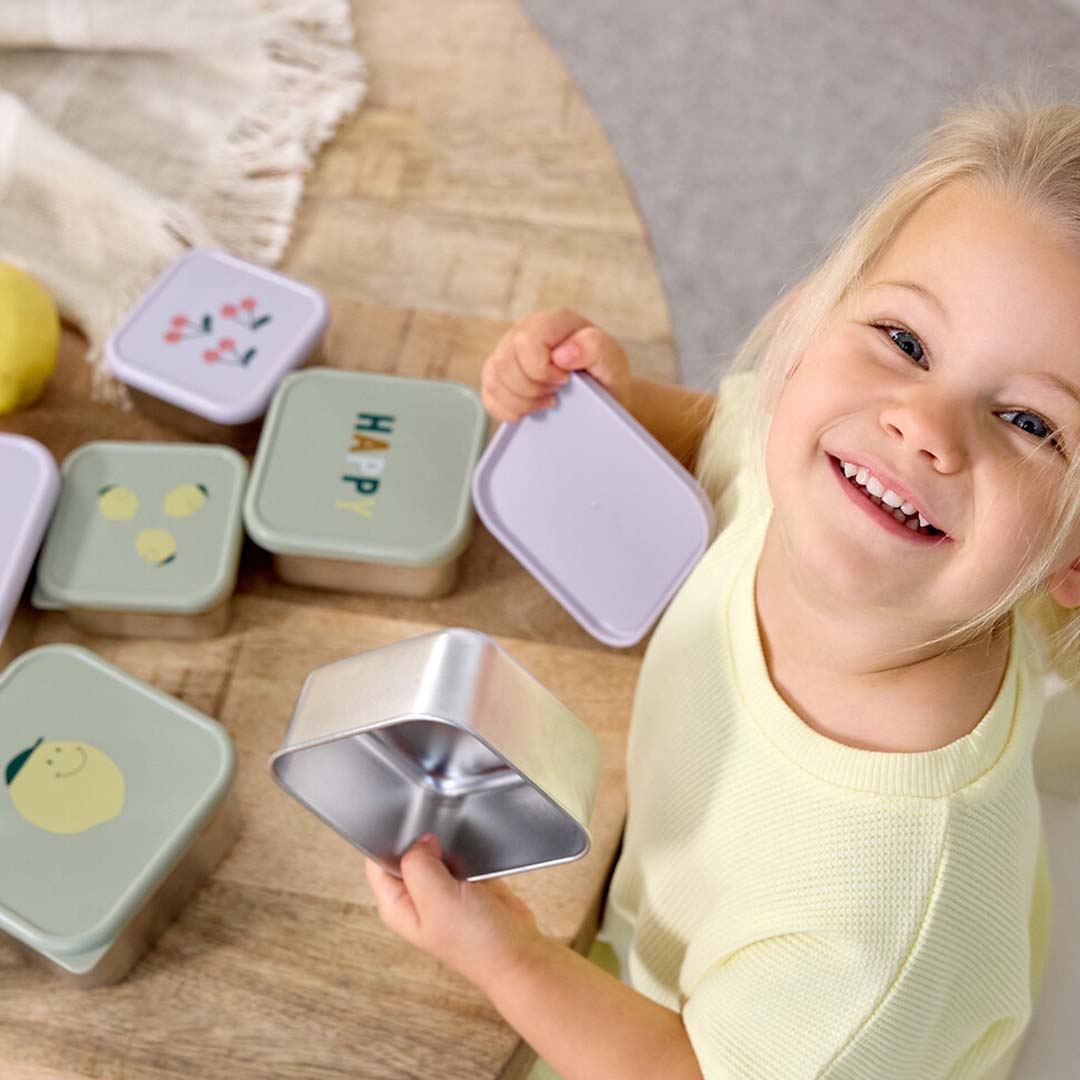 Een lachend kind houdt een open Lässig rvs snackdoosje vast, met meerdere snackdoosjes met kersen, citroenen en 'HAPPY' opdruk op een houten tafel.