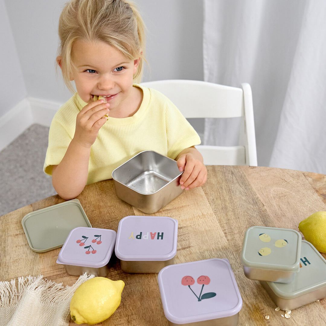 Een lachend kind eet een snack aan een houten tafel met verschillende lässig rvs snackdoosjes voor kinderen met fruit- en 'HAPPY'-opdruk.