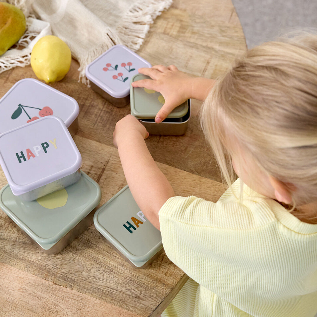Een kind pakt een lässig rvs snackdoosje voor kinderen van een houten tafel, omringd door andere doosjes met fruit- en 'HAPPY'-opdruk.