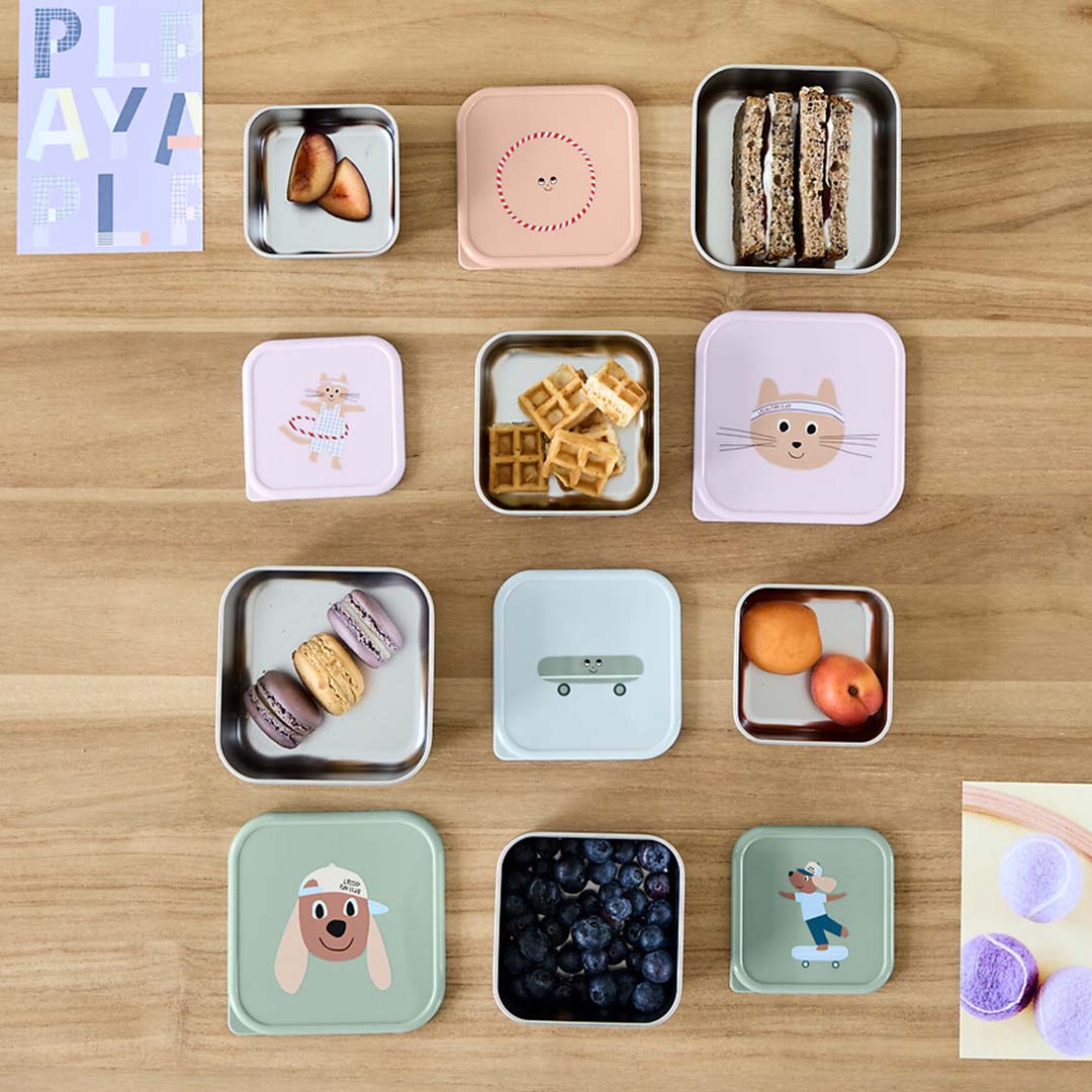 Een bovenaanzicht van diverse Lässig rvs snackdoosjes voor kinderen met dieren- en skateboardpatronen, gevuld met snacks op een houten tafel.