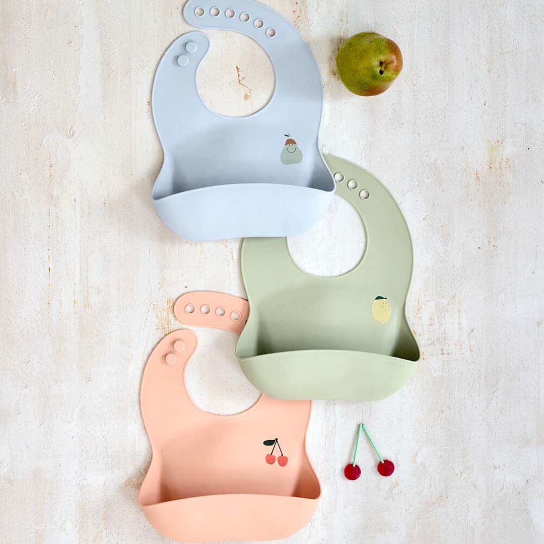 Drie lässig siliconen slabbetjes in roze, groen en blauw met fruitdesigns, naast een peer en kersen.