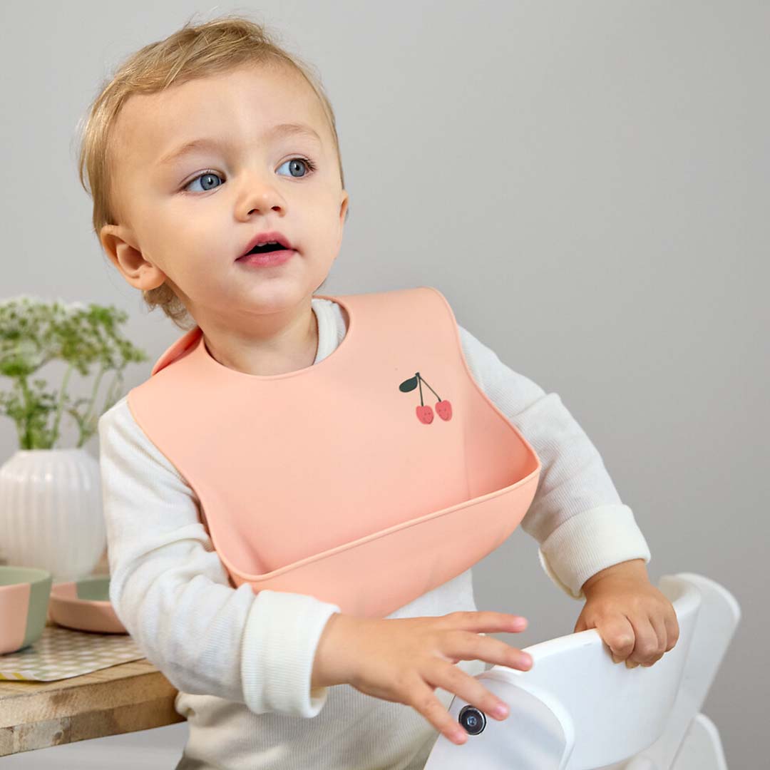 Een baby draagt het lässig roze siliconen slabbetje met kersendesign terwijl het in een kinderstoel staat.